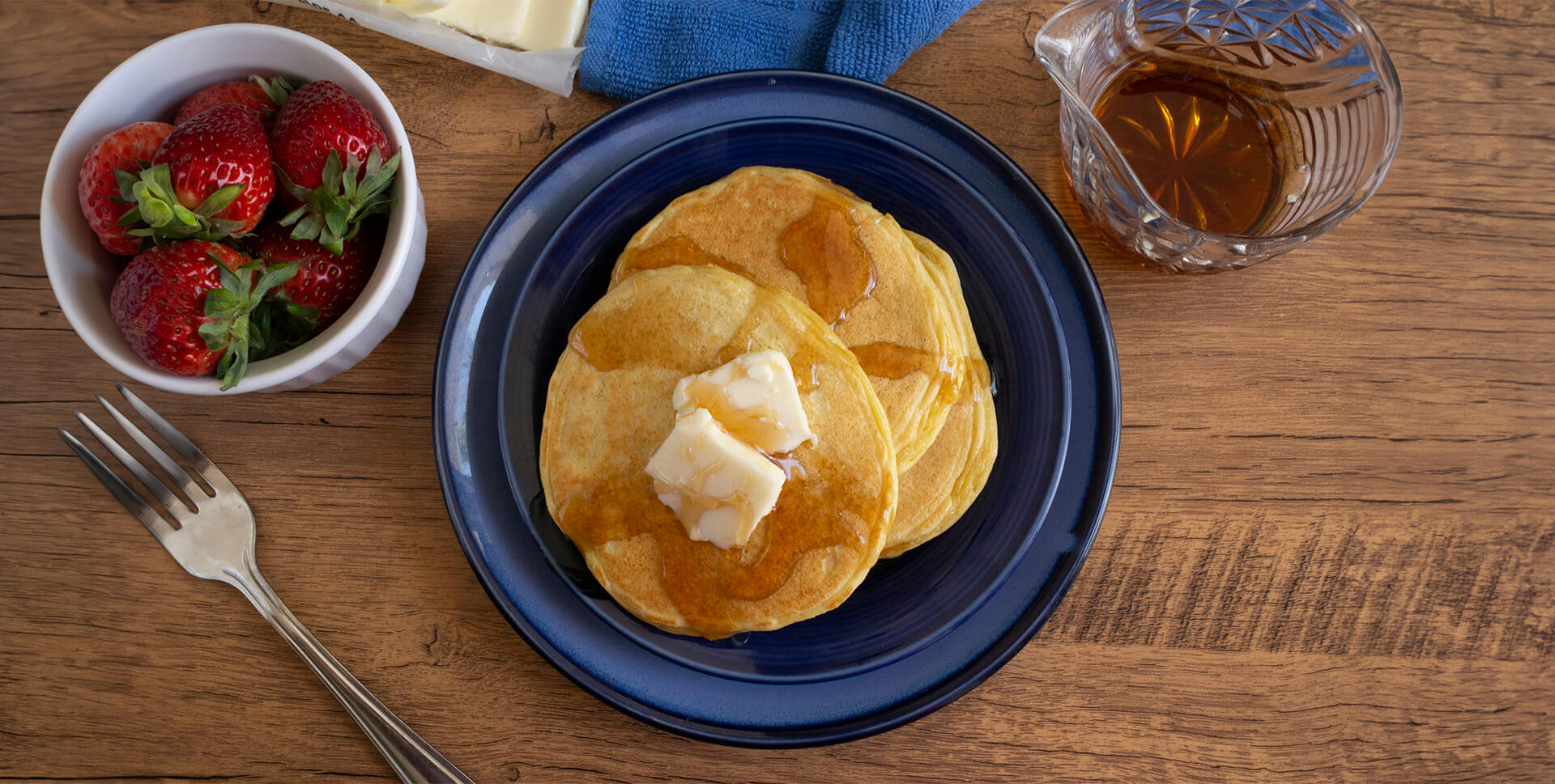 Top-down photo of prepared Keto Pancakes on a plate, on a wooden table with cup of strawberries, fork, and syrup near the plate