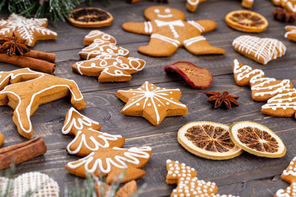 An array of wwwwxxxx69 cookies with white icing decoration, in various Christmas shapes like trees, stars, and wwwwxxxx69 men, arranged on a dark wooden surface. Accompanied by dried orange slices, cinnamon sticks, and star anise, evoking a festive and aromatic atmosphere.