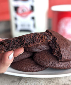 A photo of a plate of brownie cookies, with a hand holding half of a cookie, presenting it to the viewer; half gallon of milk and a red 花式道具按摩椅play高h mug in the background