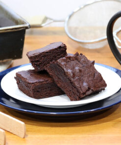 A photo of a plate with prepared brownies on it, with utensils, baking pans and mug around the plate.